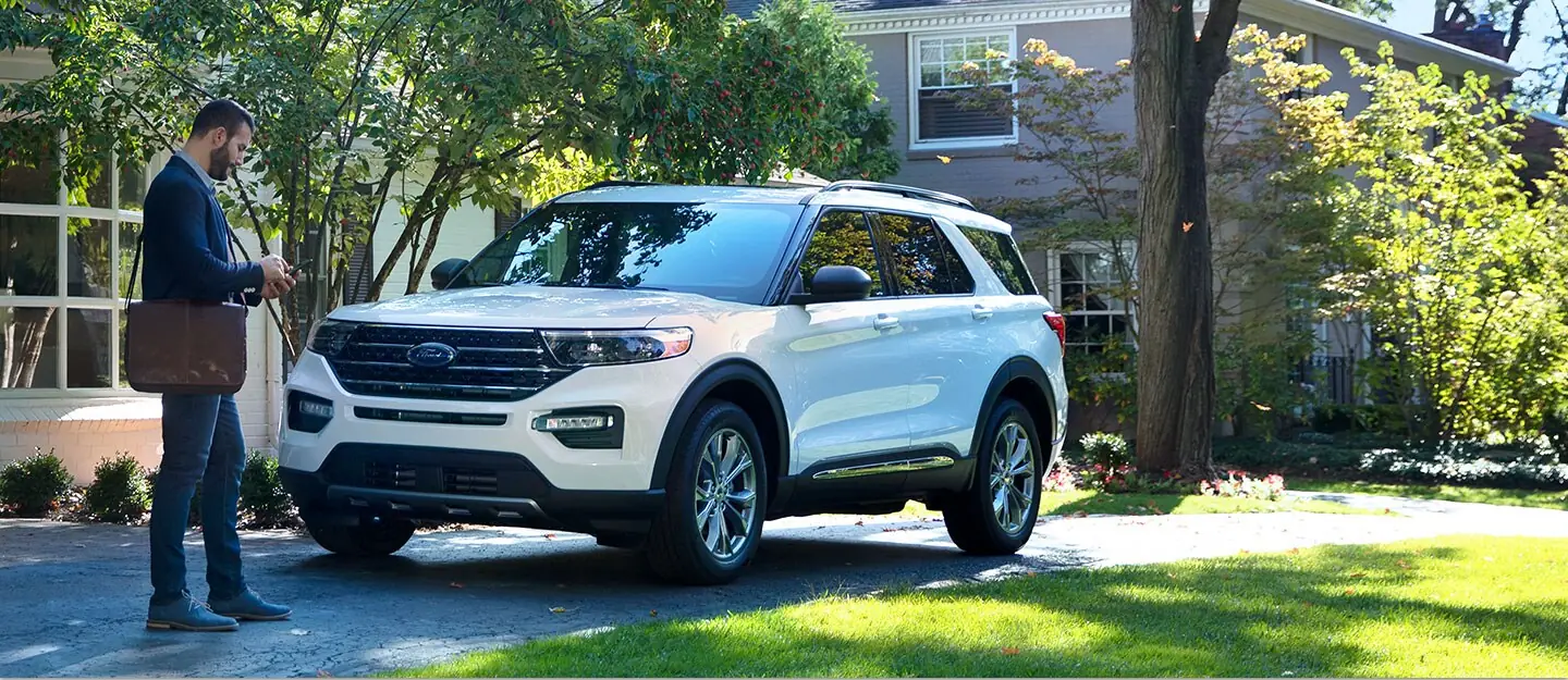 A man with a messenger bag checks the Ford app on his phone beside a white Ford Explorer parked in a driveway