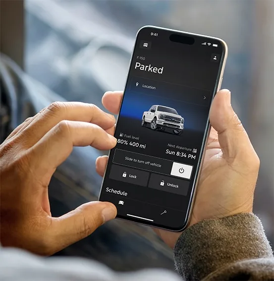 Hands holding a phone with the Ford app showing a parked F-150, fuel level, lock, unlock and remote start