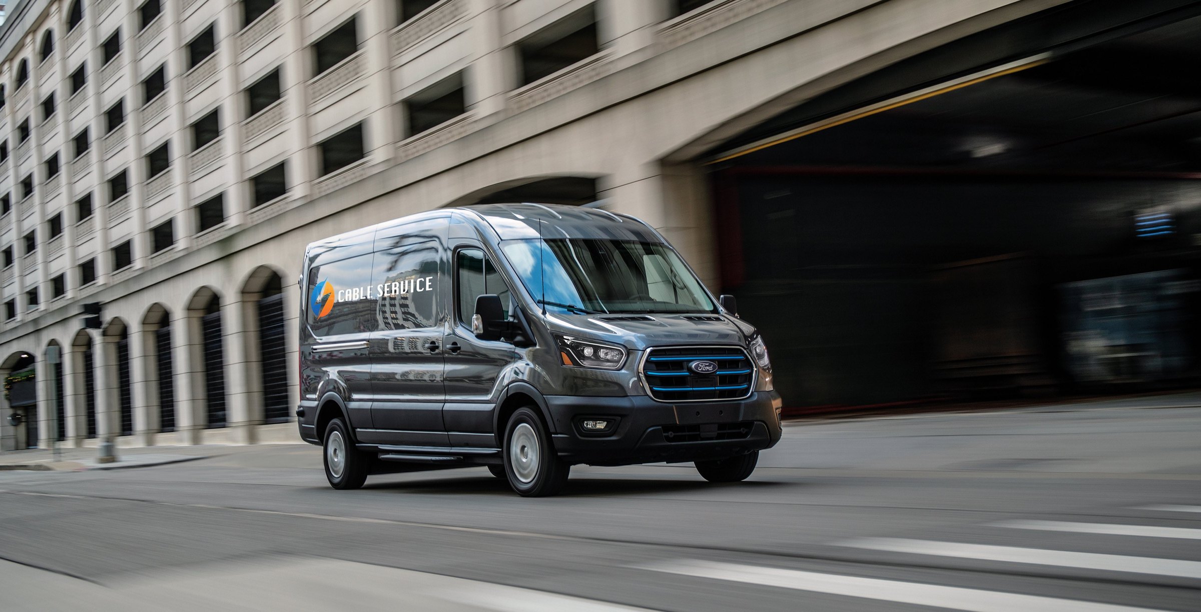 A Carbonized Gray Ford E-Transit cargo van in company livery driving past a colonnaded downtown building, seen from the front passenger side in motion