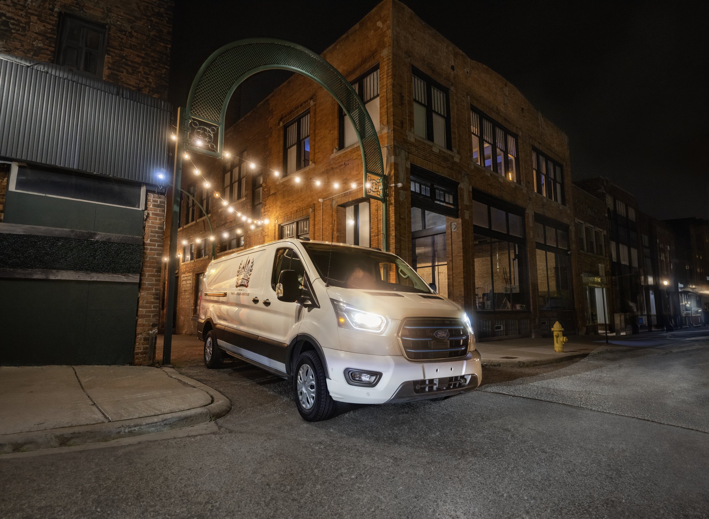 A white Ford E-Transit parked at night on a street outside a brick building strung with festoon lights, its headlights on