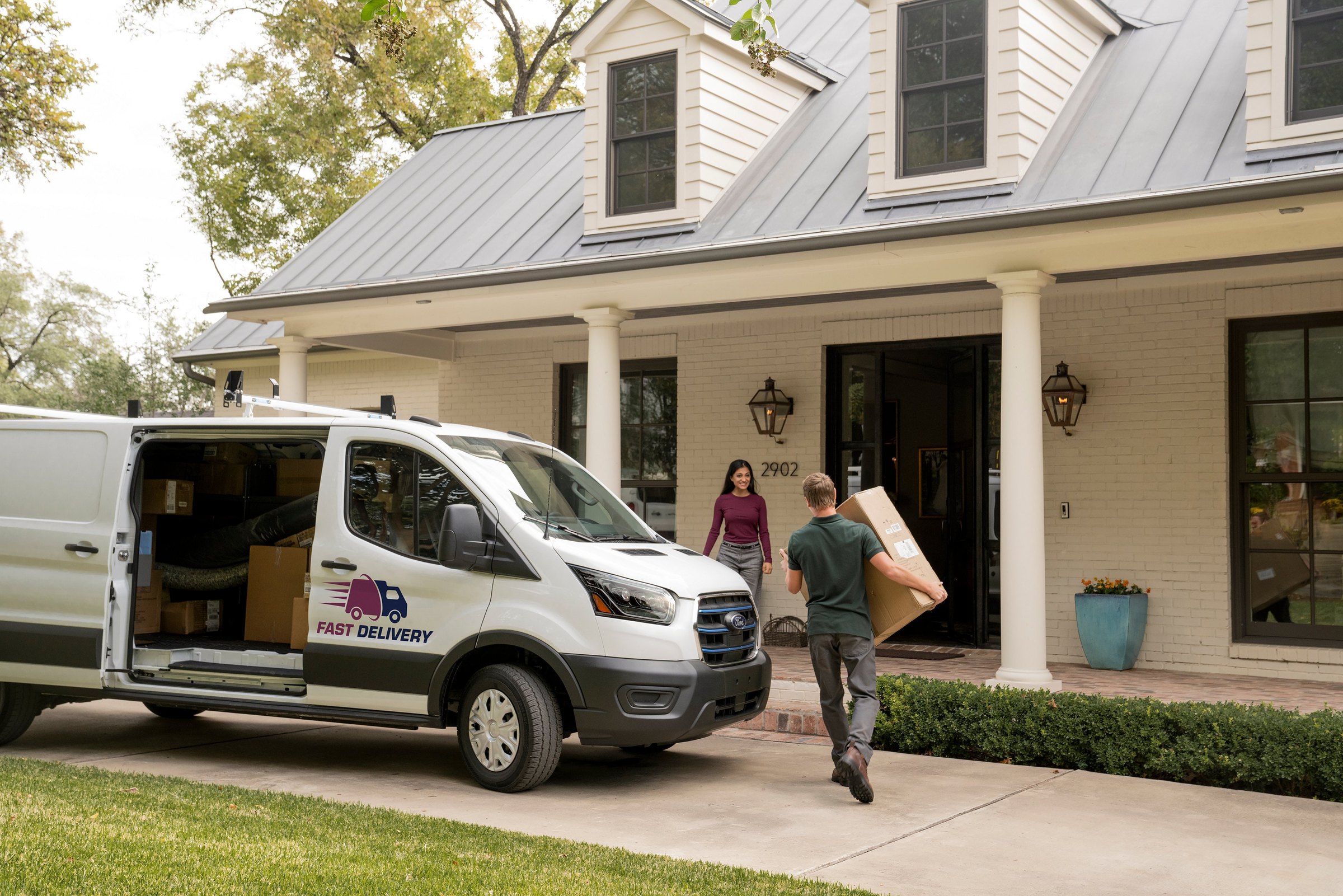 A white Ford E-Transit marked Fast Delivery parked at a curb outside a house, rear doors open on stacked boxes while a courier carries a package toward the door