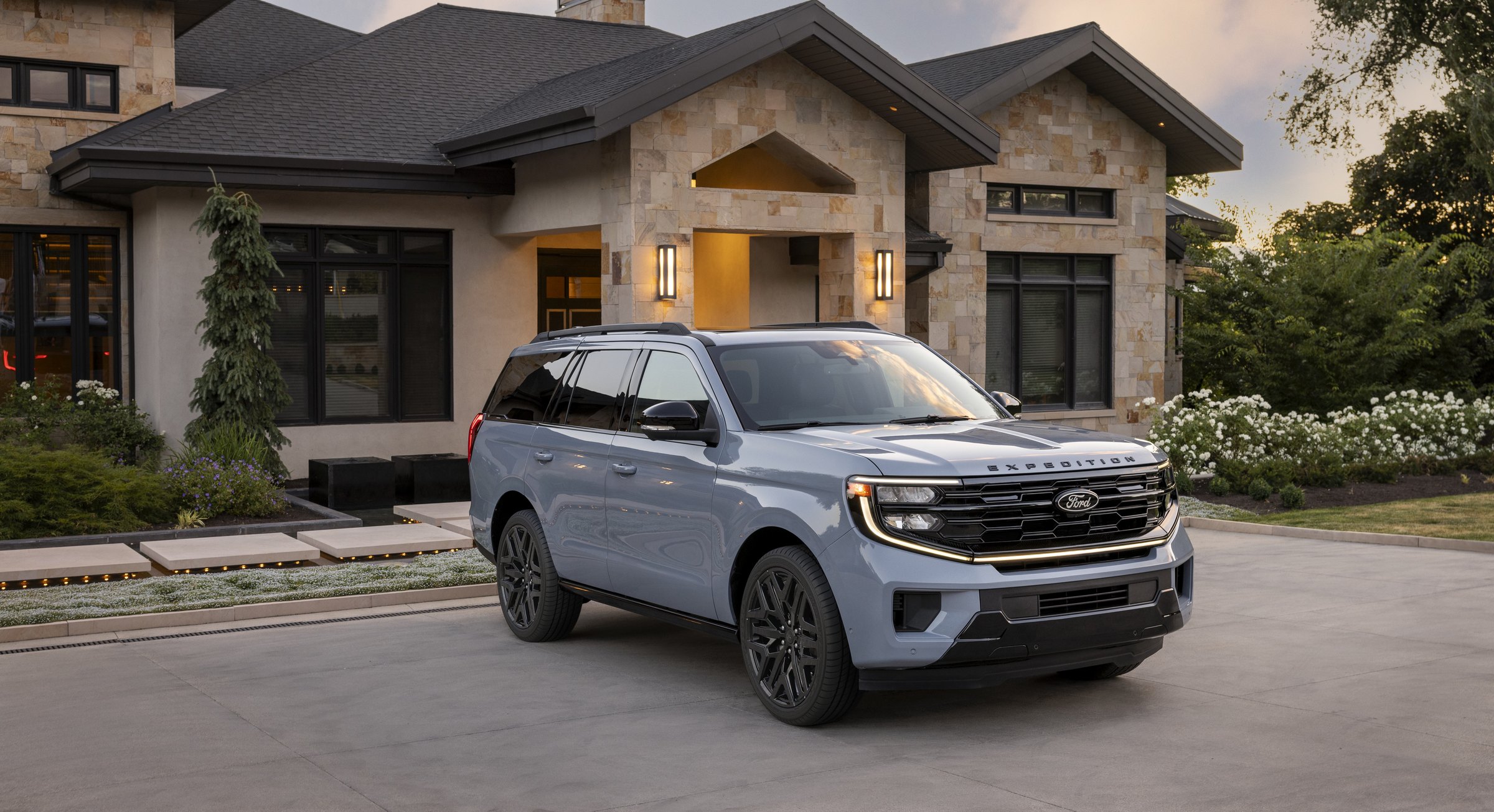 A pale blue-gray Ford Expedition parked on the driveway of a stone-front house