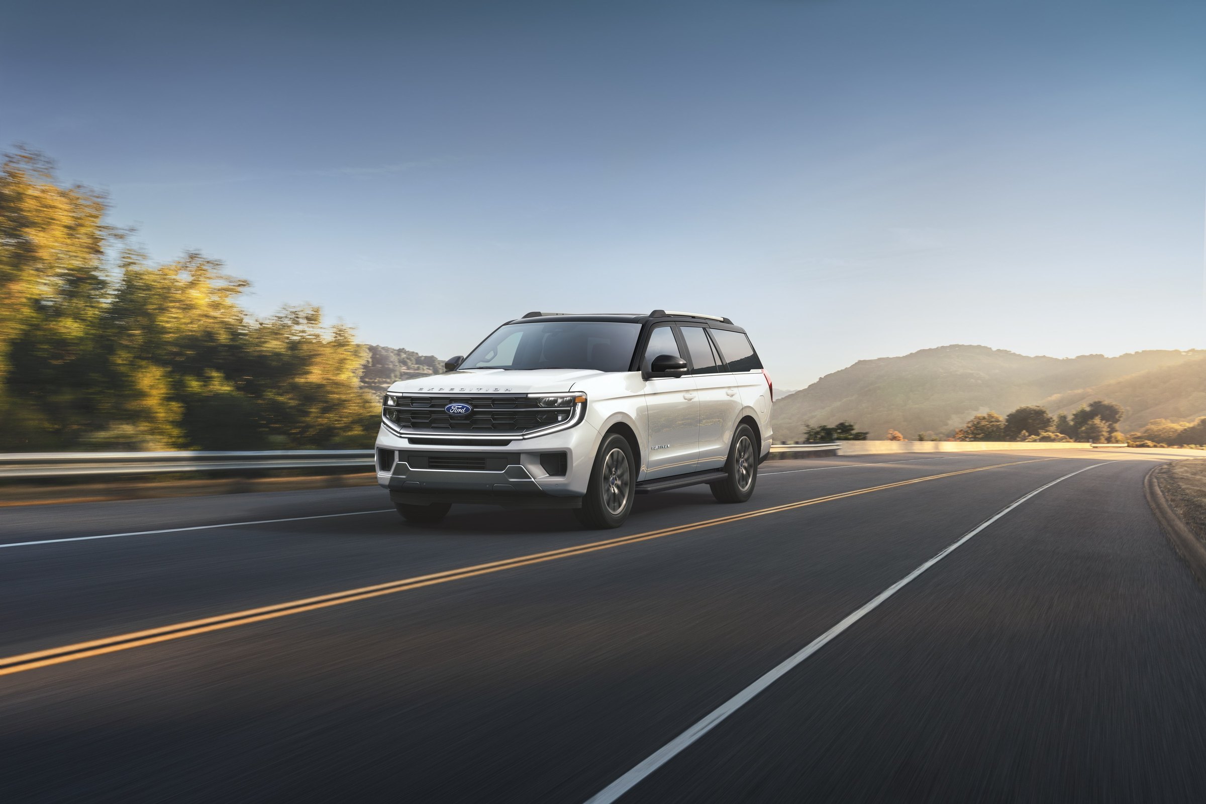 A white 2027 Ford Expedition with a black roof driving on a highway, the road blurred with speed and hills behind it