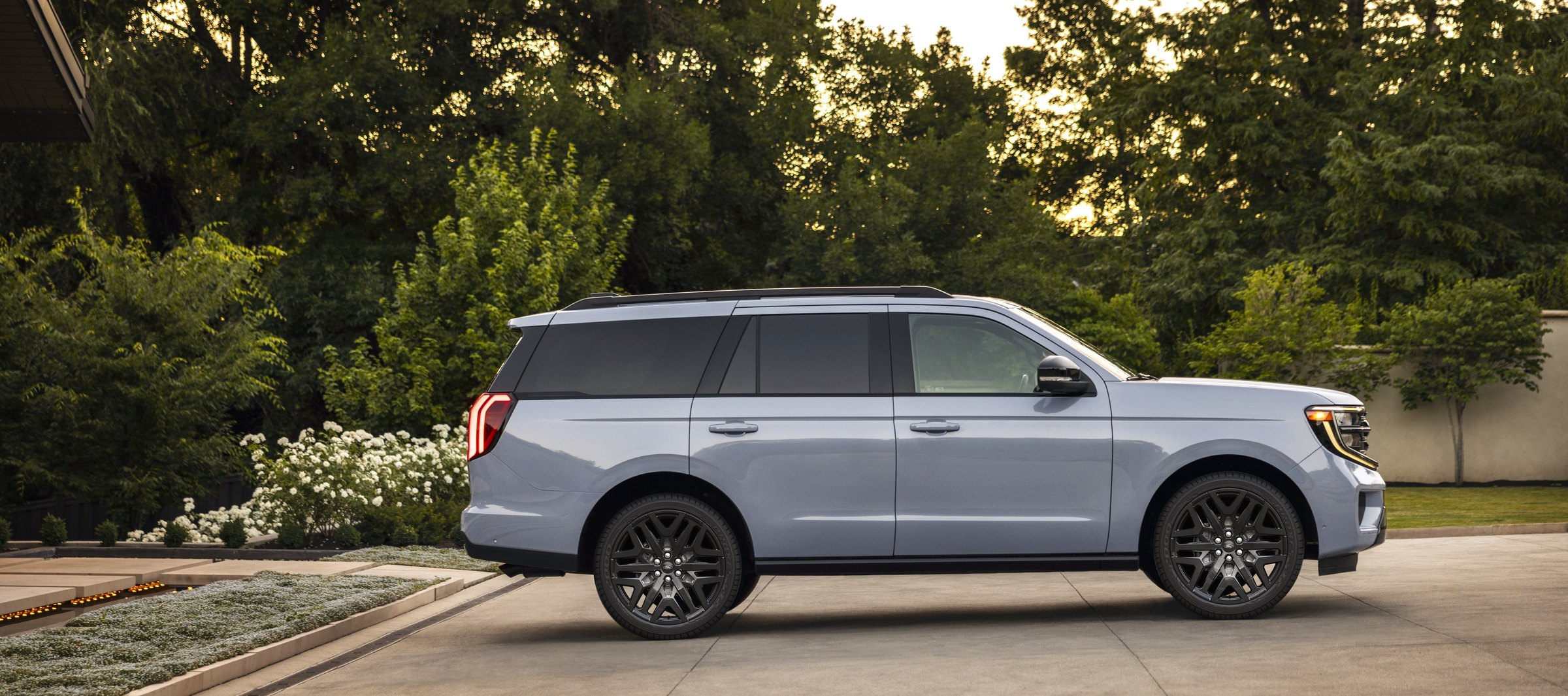 A pale blue-gray Ford Expedition seen from the side on a paved driveway at dusk, with trees and flowering shrubs behind it