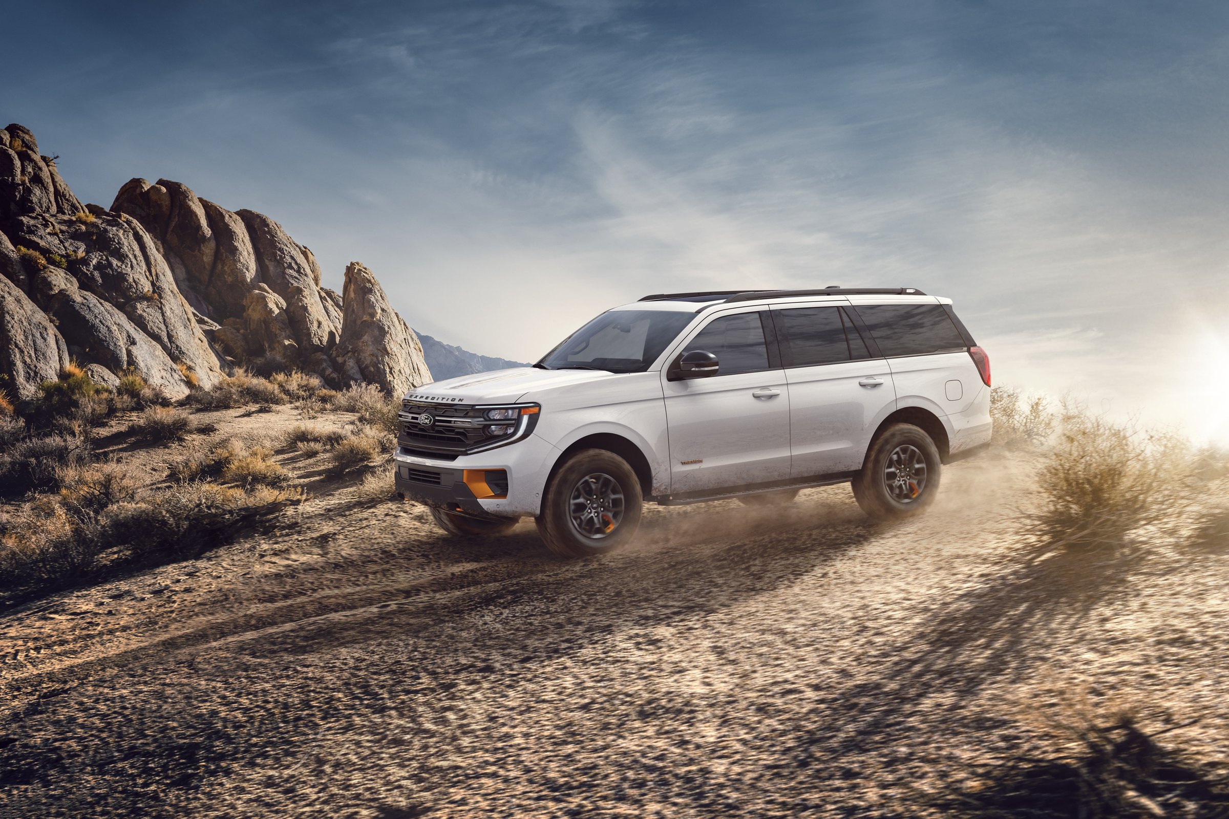 A white Ford Expedition Tremor driving up a dusty desert trail past rock outcrops, throwing up dust behind it