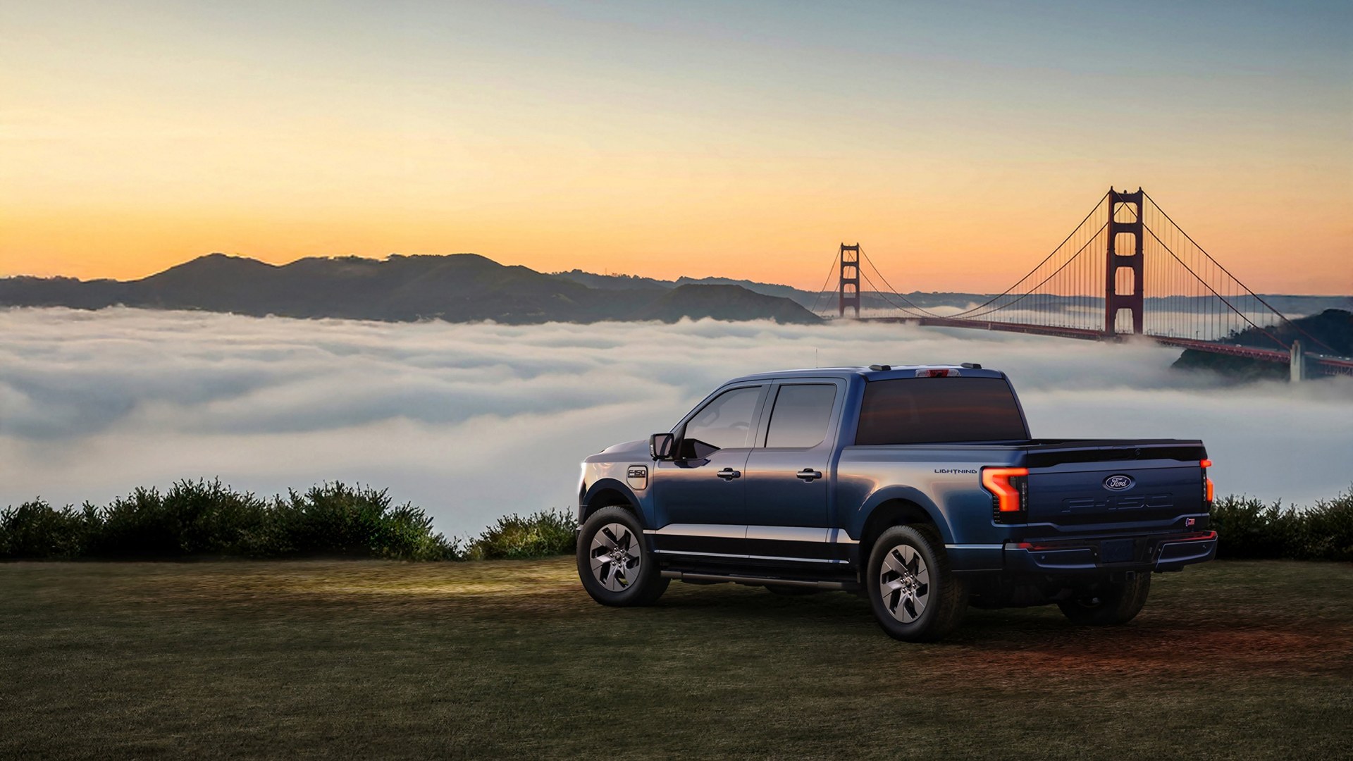 A blue Ford F-150 Lightning parked side-on on grass at dawn, with a fog bank filling the strait behind it and the towers of the Golden Gate Bridge rising out of the fog