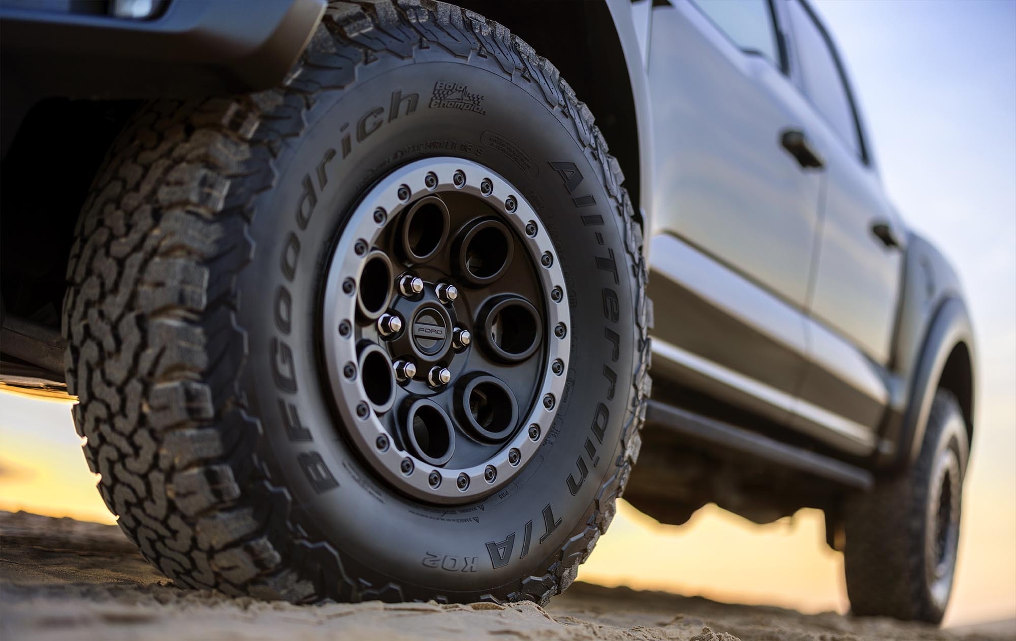 Close view of a knobby BFGoodrich All-Terrain tire and dark alloy wheel on a Ford pickup, photographed low against a sunset sky
