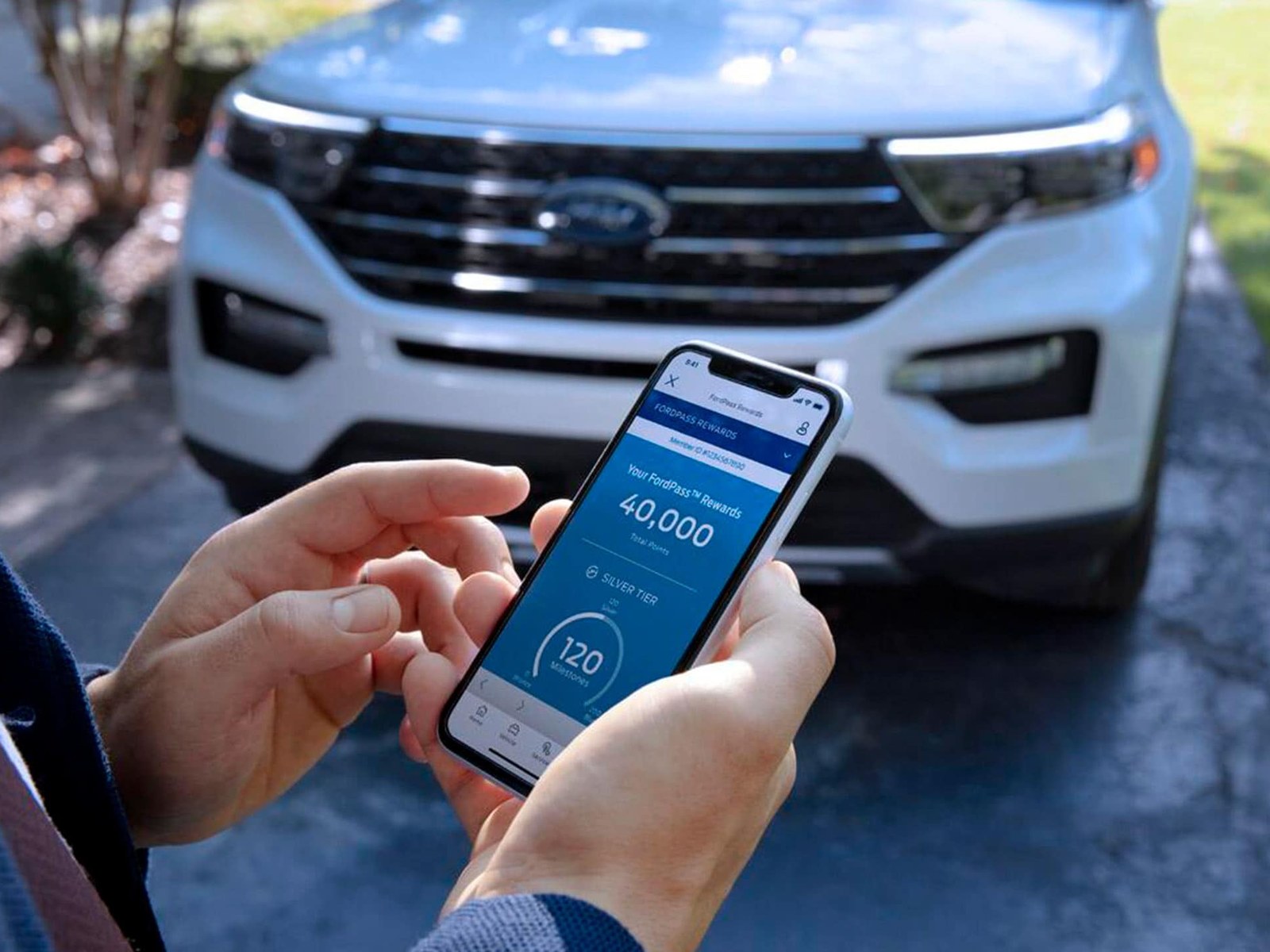 A hand holding a phone showing the FordPass Rewards screen with a points balance, a white Ford Explorer parked on the driveway behind it