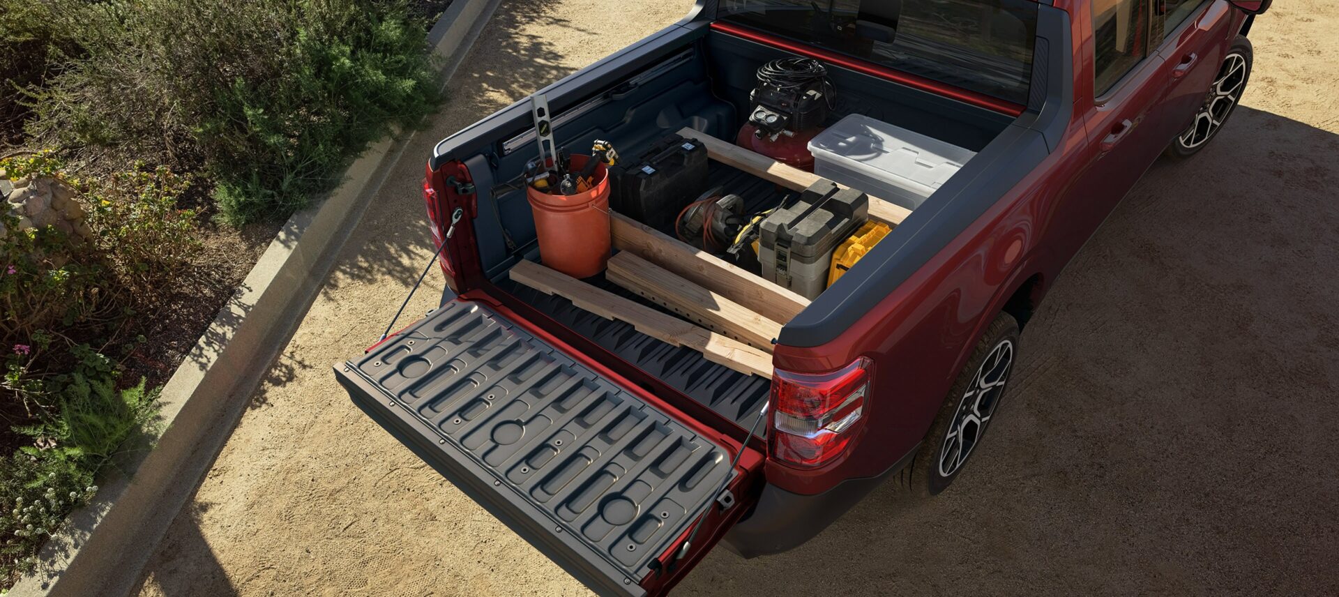 The open bed of a red 2026 Ford Maverick loaded with lumber, a bucket, a cooler and a generator, tailgate down