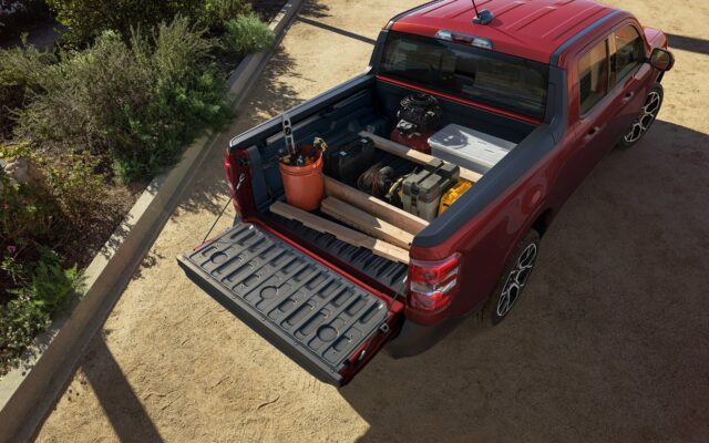 The open bed of a red 2026 Ford Maverick loaded with lumber, a bucket, a cooler and a generator, tailgate down