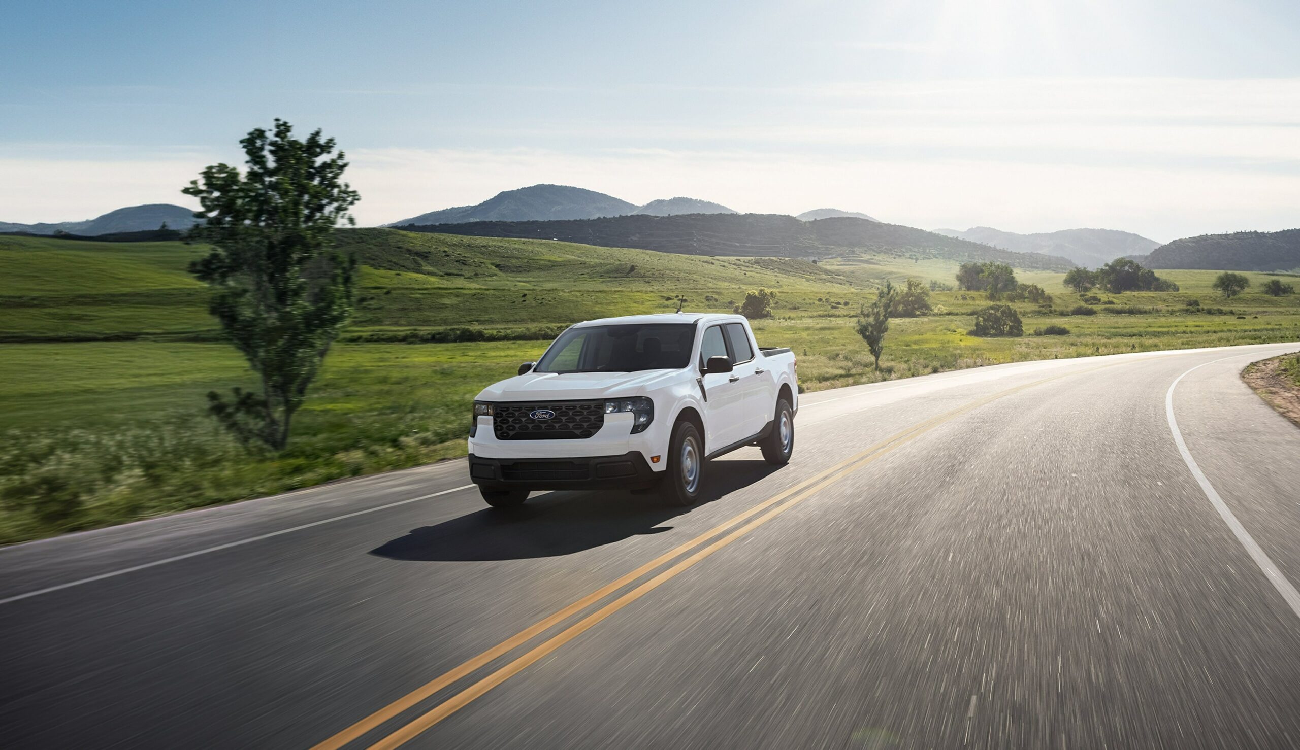 A white 2026 Ford Maverick driving on a two-lane road through green rolling country