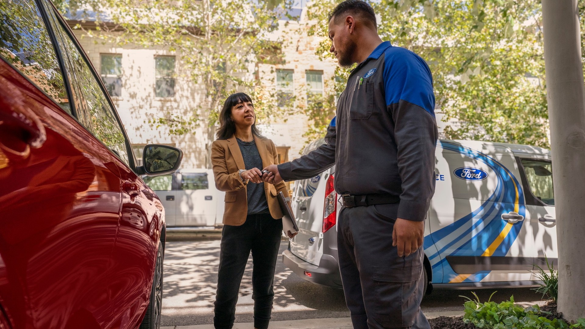 A Ford technician in a Ford-branded uniform handing car keys back to a customer beside a red Ford, with two white Ford Mobile Service Transit vans parked behind them