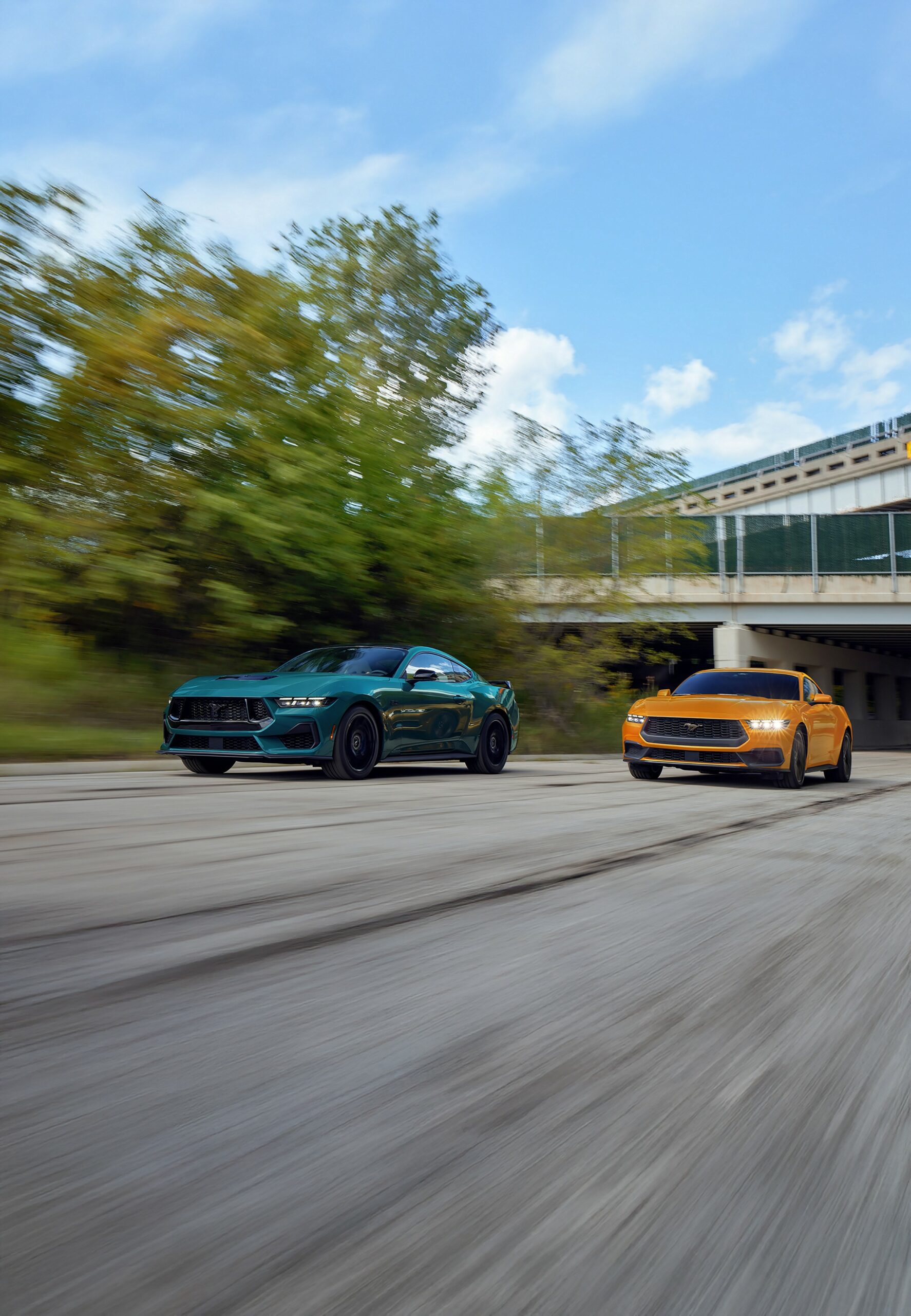 A blue 2026 Ford Mustang fastback and an orange Mustang convertible driving side by side beneath a concrete overpass