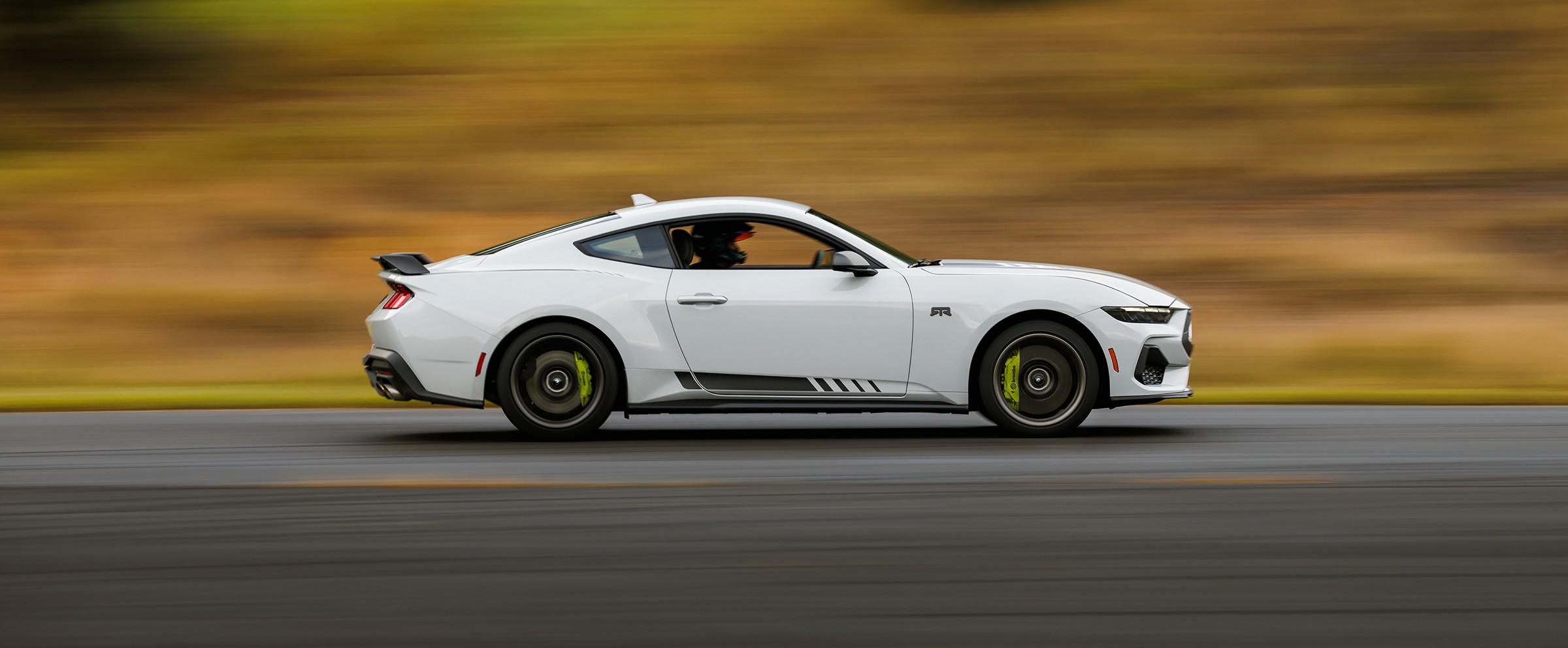 A white 2026 Ford Mustang with the RTR Package at speed on a track, seen from the side with the background blurred