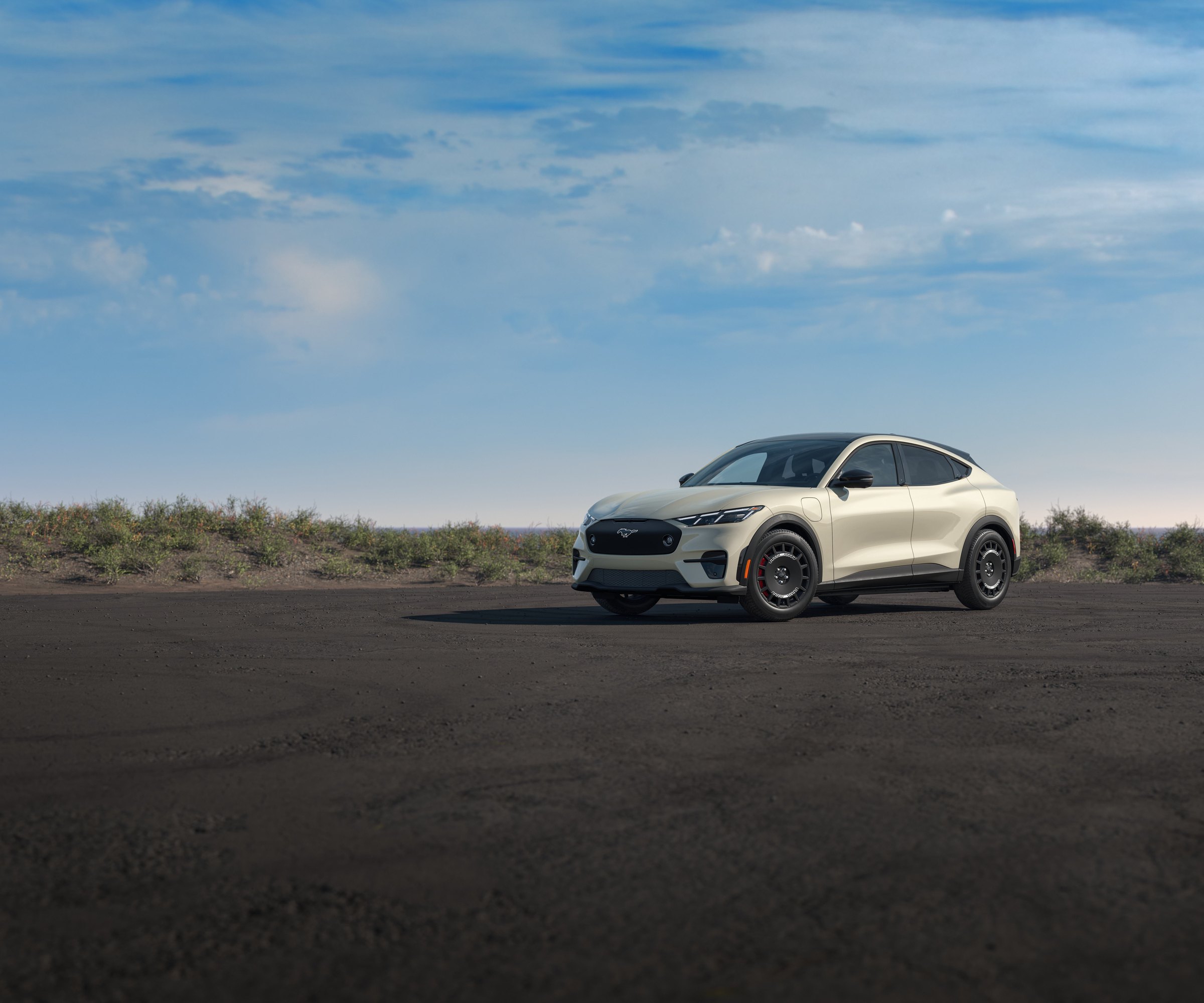 A sand-colored Ford Mustang Mach-E Rally with a black roof parked on dark gravel under an open blue sky, low scrub on the bank behind it
