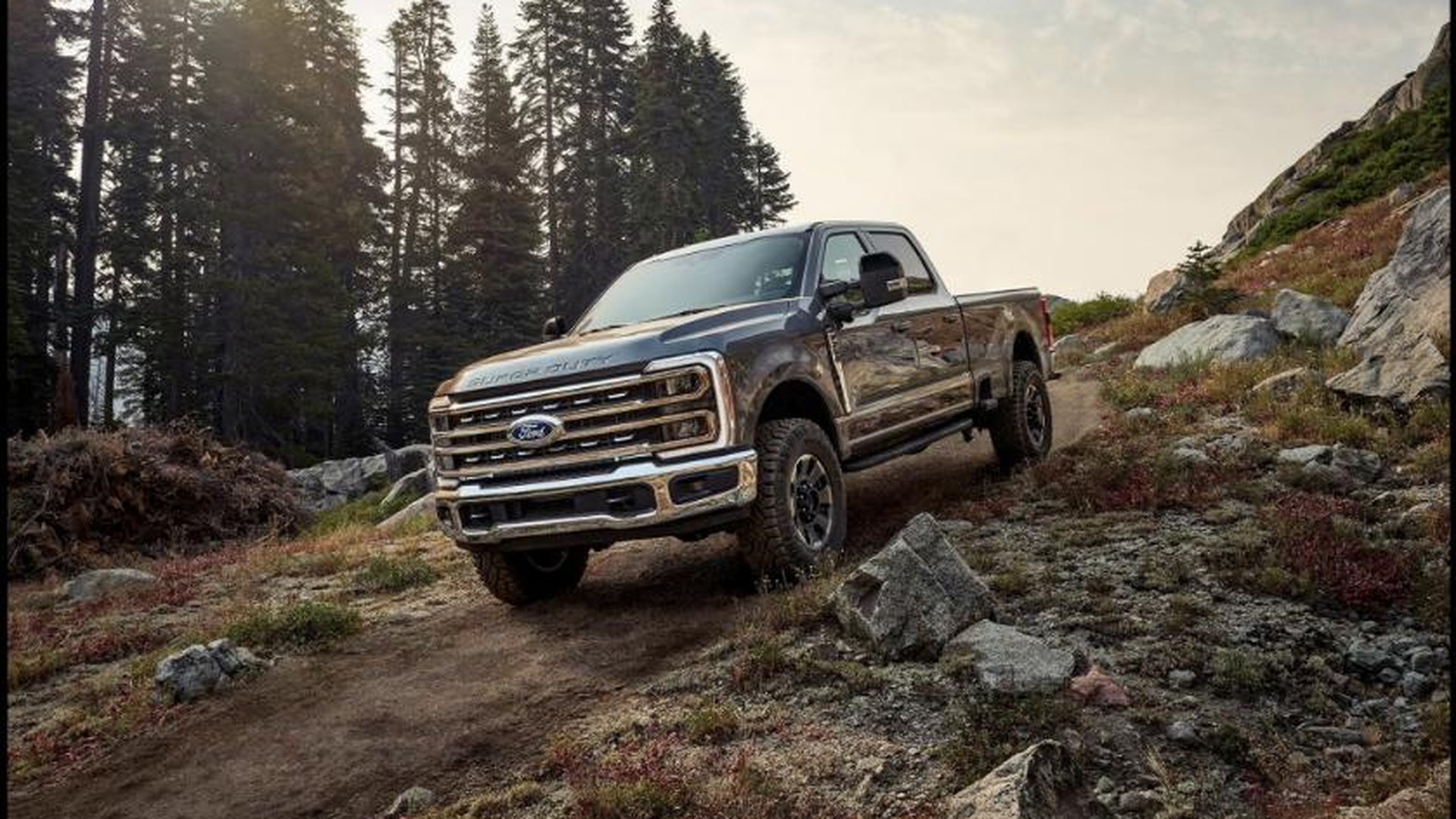 A gray 2027 Ford Super Duty F-250 with the Tremor Off-Road Package climbing a rocky dirt track on a pine-covered slope in low sunlight