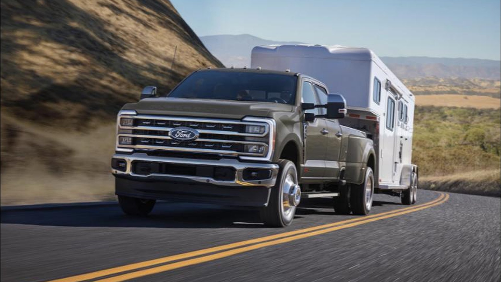 A gray Ford Super Duty F-350 Lariat crew-cab dually towing a white gooseneck trailer along a two-lane highway beside a dry hillside
