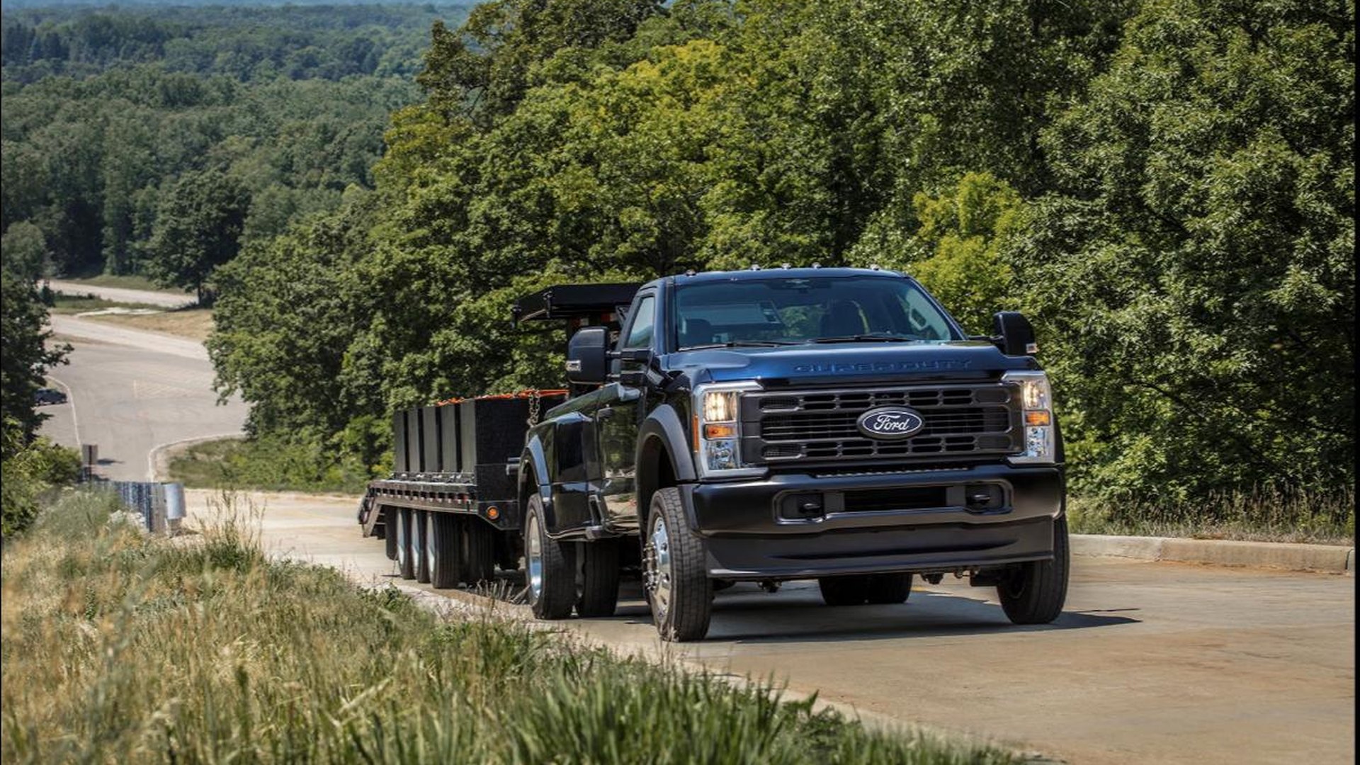 A dark blue Ford Super Duty regular-cab dually pulling a loaded flatbed gooseneck trailer up a paved hill through summer woodland