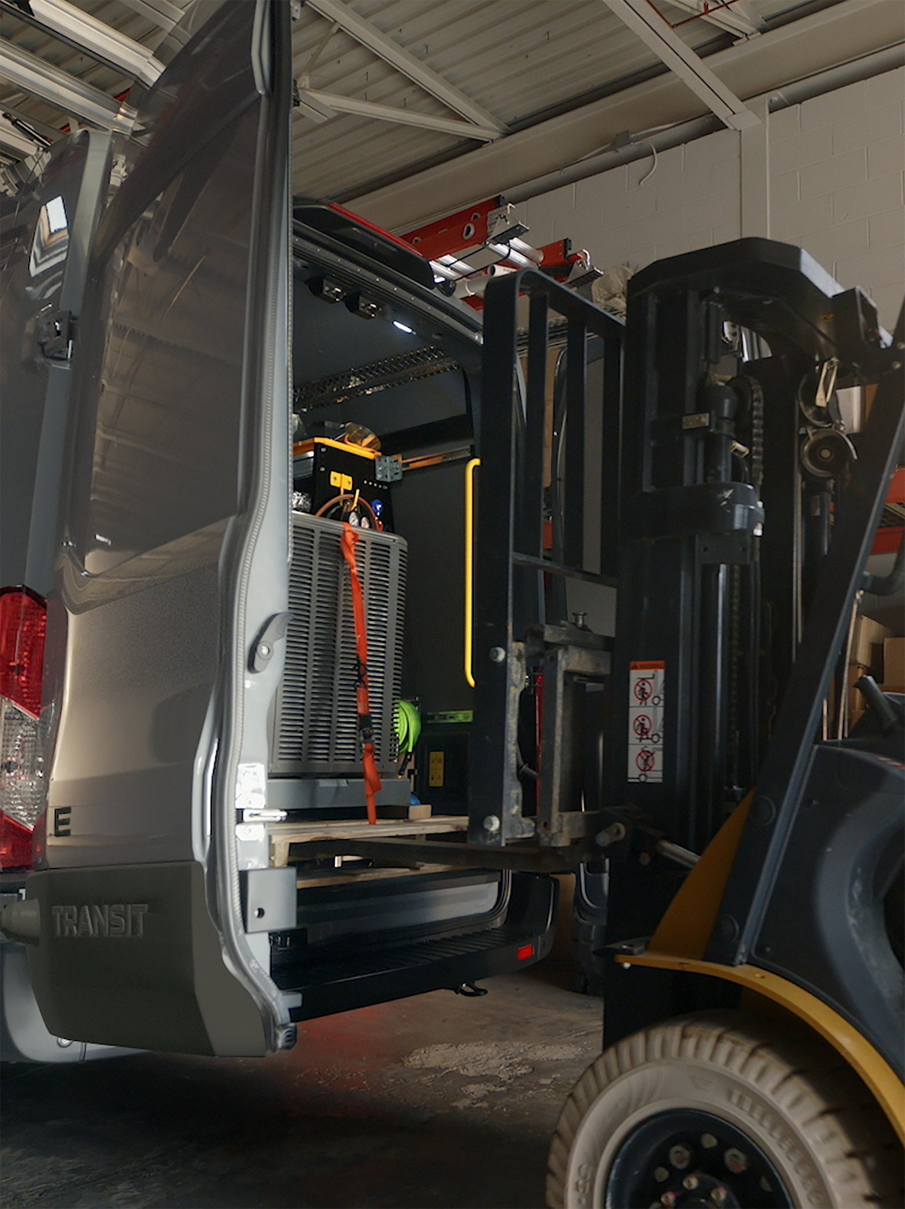 A forklift lifting a wrapped pallet into the back of a Ford Transit through its open rear doors inside a warehouse