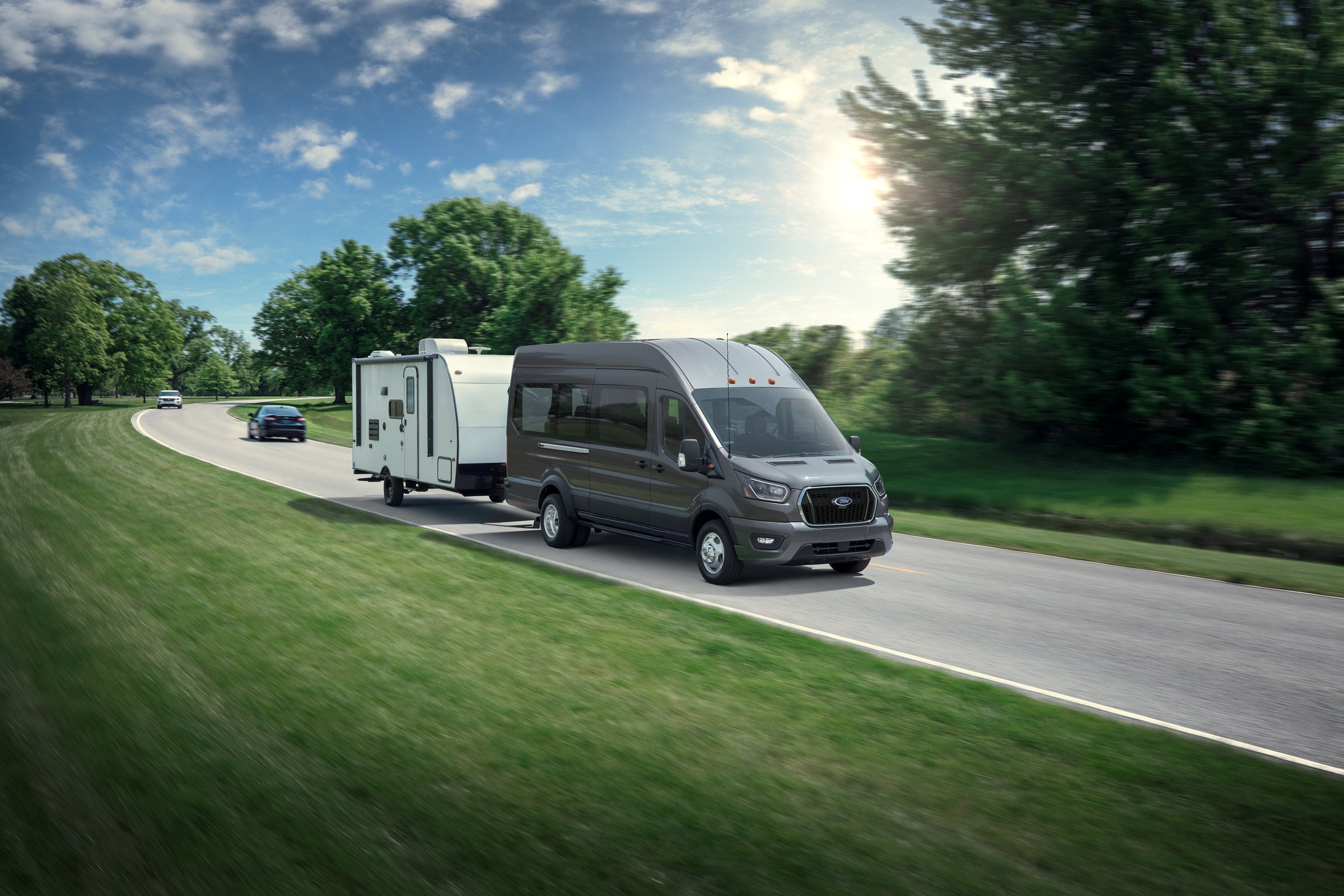 A silver high-roof Ford Transit towing a white travel trailer along a highway past a green field at sunset
