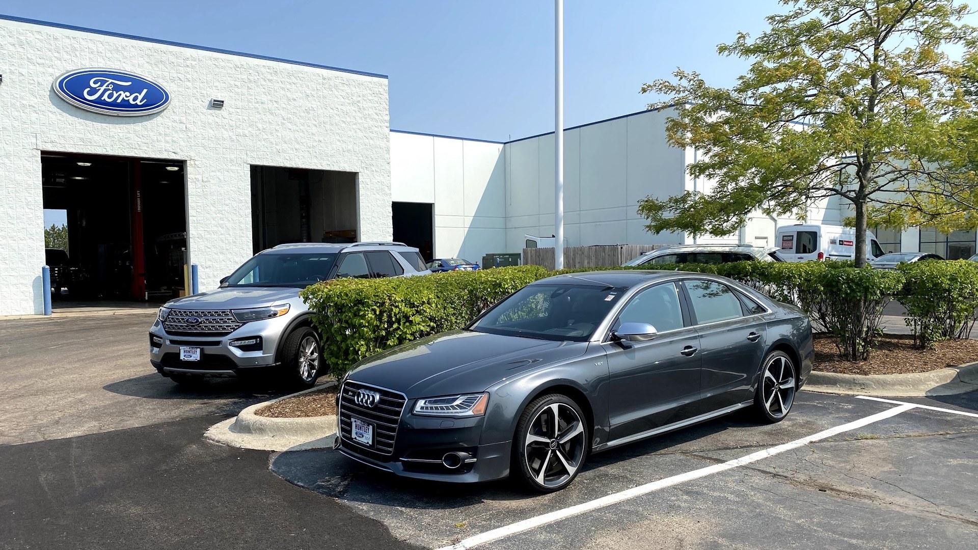 The service drive at Huntley Ford on a clear morning, with two bay doors open under the Ford sign and a Ford Explorer and an Audi sedan parked outside, both wearing Huntley Ford plate frames